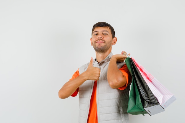 Varón joven en camiseta, chaqueta sosteniendo bolsas de compras, mostrando el pulgar hacia arriba y mirando alegre, vista frontal.