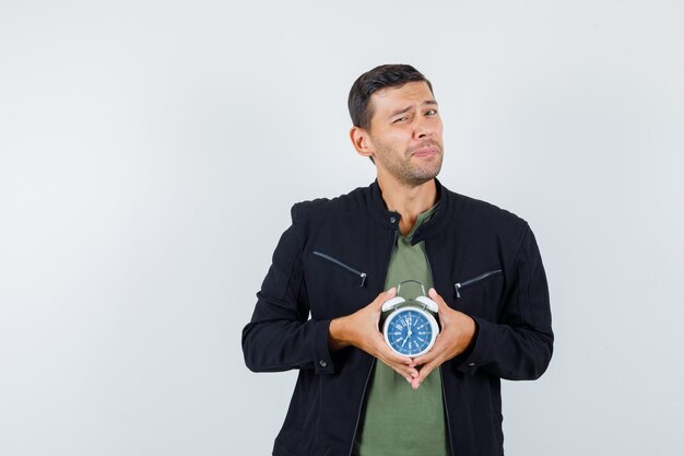 Varón joven en camiseta, chaqueta sosteniendo reloj despertador y mirando triste, vista frontal.