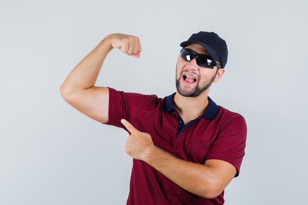 Varón joven en camiseta, gafas de sol apuntando a los músculos de su brazo y mirando alegre, vista frontal.