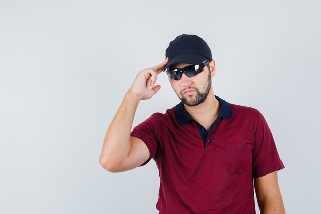 Varón joven en camiseta, gafas de sol de pie mientras endurece su gorra y mira seria, vista frontal.