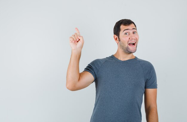 Varón joven en camiseta gris apuntando hacia arriba y mirando feliz