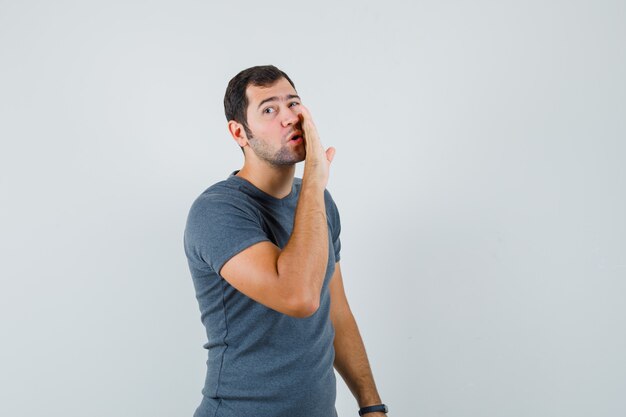 Varón joven en camiseta gris contando secreto detrás de la mano