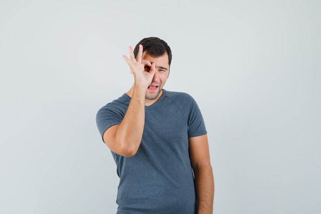 Varón joven en camiseta gris mostrando signo ok en el ojo y mirando curioso