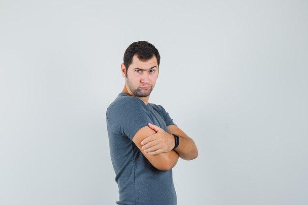Varón joven en camiseta gris de pie con los brazos cruzados y mirando dudoso
