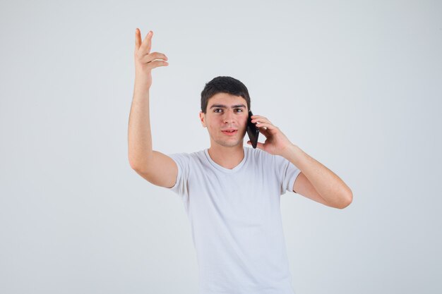 Varón joven en camiseta hablando por teléfono móvil mientras levanta el brazo y mira enfocado, vista frontal.