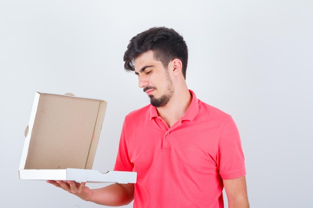 Varón joven en camiseta mirando la caja de pizza abierta y mirando vacilante, vista frontal.
