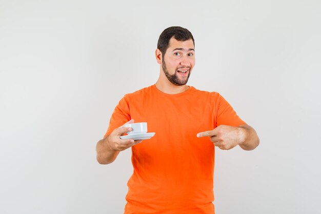 Varón joven en camiseta naranja apuntando a la taza con platillo y mirando alegre, vista frontal.