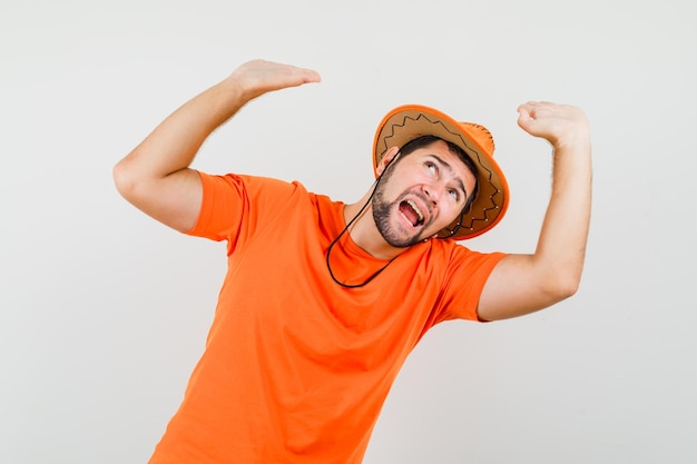 Varón joven en camiseta naranja, sombrero levantando las palmas para defenderse y mirando asustado, vista frontal.