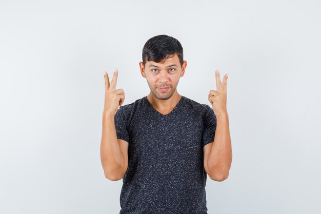 Varón joven en camiseta negra que muestra v-sign y mirando contento, vista frontal.