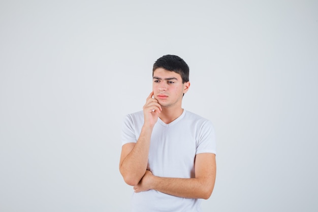 Varón joven en camiseta de pie en pose de pensamiento y mirando pensativo, vista frontal.