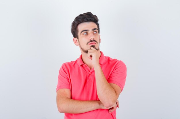 Varón joven en camiseta de pie en pose de pensamiento y mirando sensible, vista frontal.