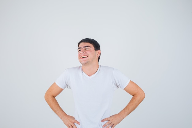 Varón joven en camiseta posando con las manos en la cintura y mirando alegre, vista frontal.