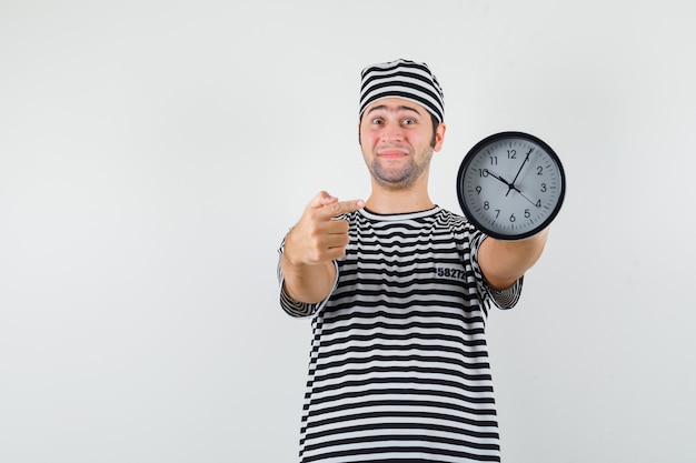 Varón joven en camiseta a rayas, gorra apuntando al reloj y mirando contento, vista frontal.