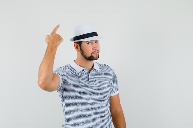 Varón joven en camiseta, sombrero apuntando hacia afuera con el dedo hacia arriba y mirando serio, vista frontal.