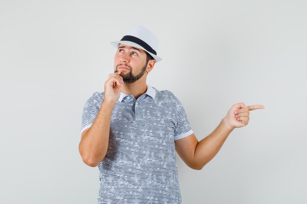 Varón joven en camiseta, sombrero apuntando hacia un lado mientras mira hacia arriba y mira soñadora, vista frontal.