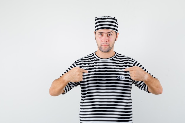 Varón joven en camiseta, sombrero apuntando a sí mismo y mirando orgulloso, vista frontal.