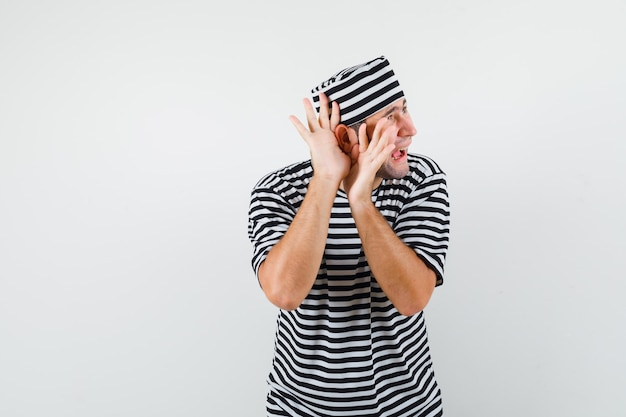 Foto gratuita varón joven en camiseta, sombrero cogidos de la mano cerca de la oreja y mirando confundido, vista frontal.