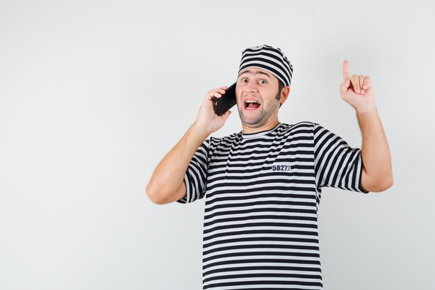 Varón joven en camiseta, sombrero hablando por teléfono móvil, apuntando hacia arriba y mirando feliz, vista frontal.