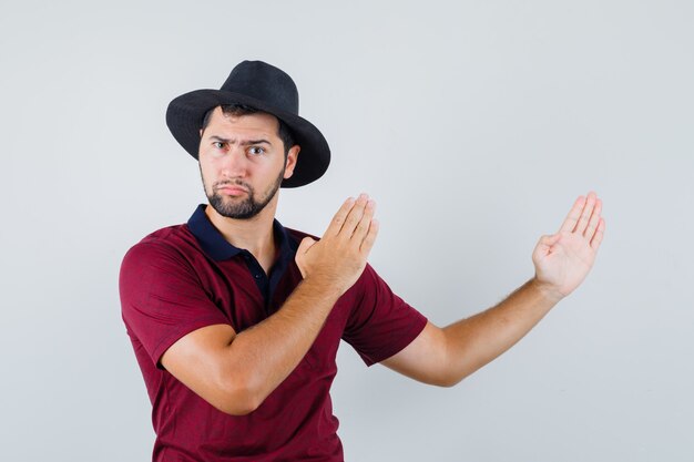 Varón joven en camiseta, sombrero mostrando gesto de tajo de karate y mirando enojado, vista frontal.