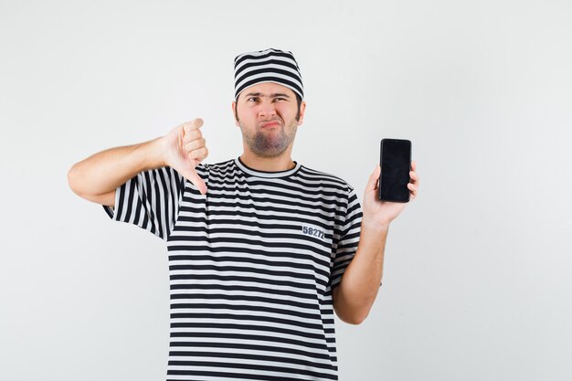 Varón joven en camiseta, sombrero sosteniendo teléfono móvil, mostrando el pulgar hacia abajo y mirando disgustado, vista frontal.