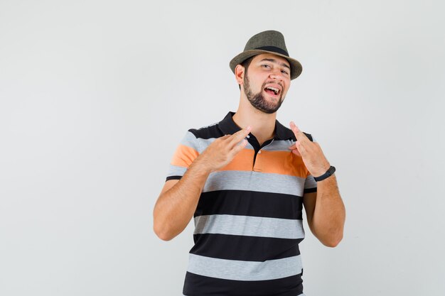 Varón joven en camiseta, sombrero tirando de su camiseta con los dedos y mirando orgulloso, vista frontal.