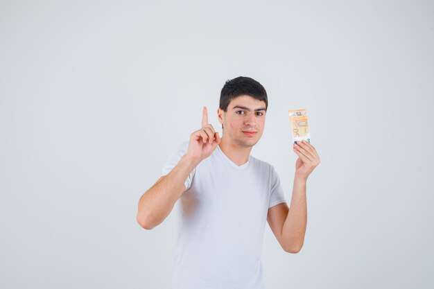 Varón joven en camiseta sosteniendo eurobanknote, apuntando hacia arriba y mirando confiado, vista frontal.