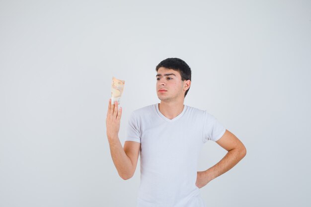 Varón joven en camiseta sosteniendo eurobanknote, mirando eurobanknote y mirando enfocado, vista frontal.