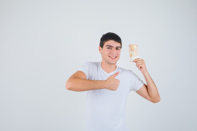 Varón joven en camiseta sosteniendo eurobanknote, mostrando el pulgar hacia arriba y mirando complacido, vista frontal.