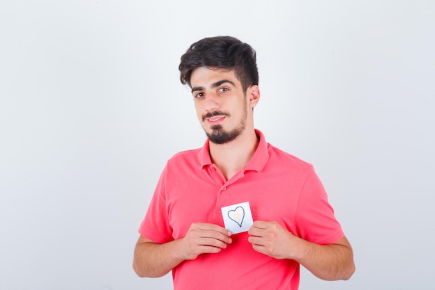 Varón joven en camiseta sosteniendo una nota adhesiva y mirando bonita, vista frontal.