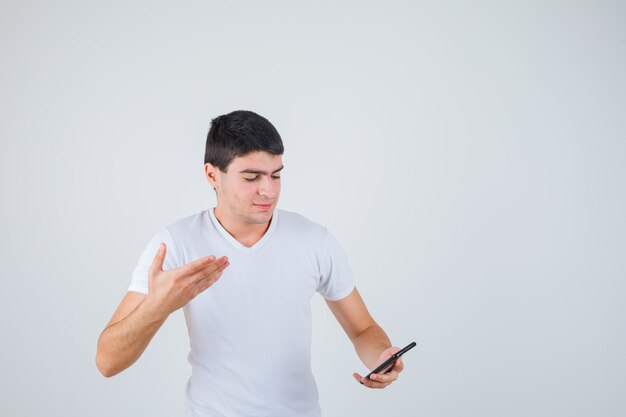 Varón joven en camiseta sosteniendo el teléfono mientras levanta la mano y mira enfocado, vista frontal.
