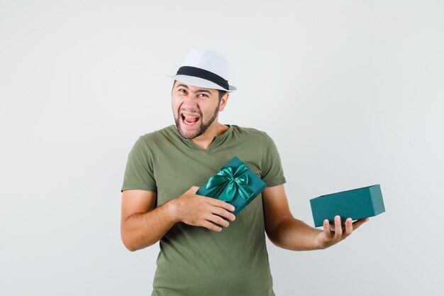 Varón joven en camiseta verde y sombrero sosteniendo caja presente abierta y mirando dichoso