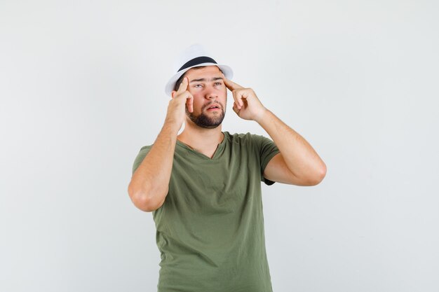 Varón joven en camiseta verde y sombrero sosteniendo los dedos en las sienes y mirando agotado