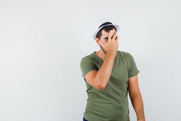 Foto gratuita varón joven en camiseta verde y sombrero sosteniendo la mano en la cara y mirando asustado, vista frontal.