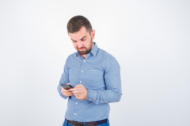 Varón joven charlando por teléfono móvil en camisa, jeans y mirando vacilante. vista frontal.