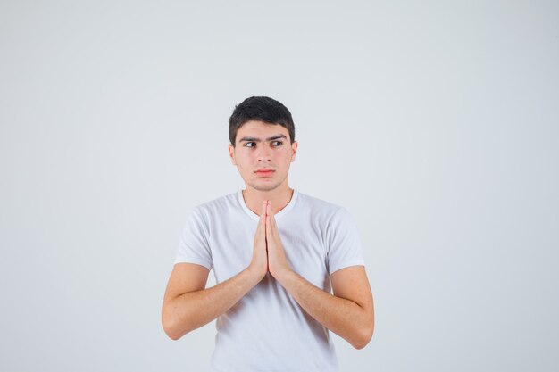 Varón joven cogidos de la mano en gesto de oración en camiseta y mirando esperanzado. vista frontal.