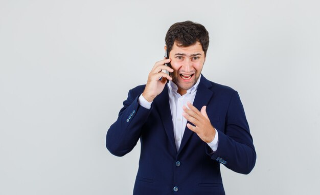 Varón joven enojado mientras habla por teléfono inteligente en traje, vista frontal.