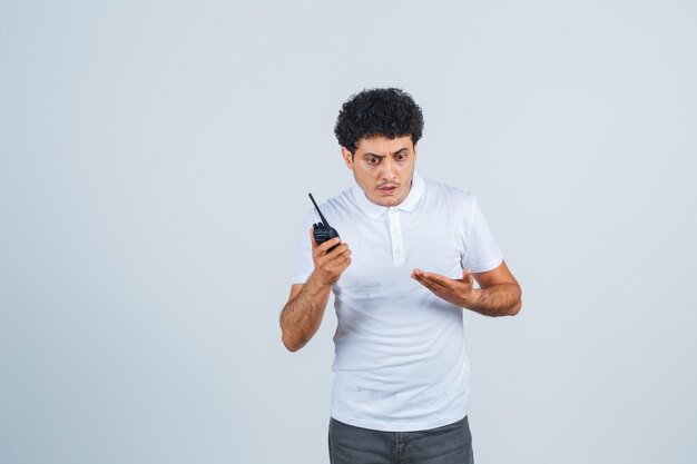 Varón joven escuchando el teléfono walkie talkie en camiseta blanca, pantalones y mirando desconcertado. vista frontal.