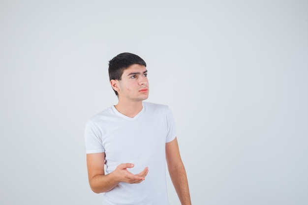 Varón joven estirando la mano en gesto de interrogación en camiseta y mirando pensativo, vista frontal.