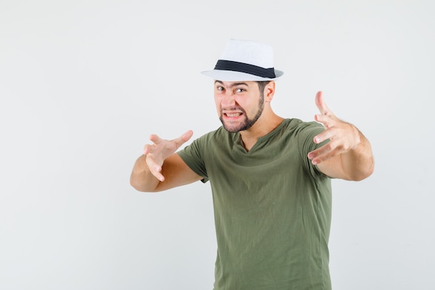 Varón joven estirando las manos como agarrando algo en camiseta verde y sombrero y luciendo agresivo