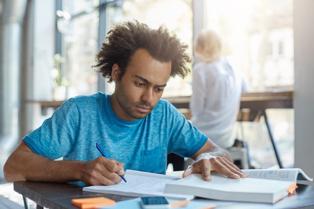 Varón joven estudiante concentrado en camiseta azul sentado en el escritorio en el interior reescribiendo información del libro en el libro de copia. Atractivo hombre de piel oscura escribiendo sinopsis mientras está sentado en la acogedora cafetería