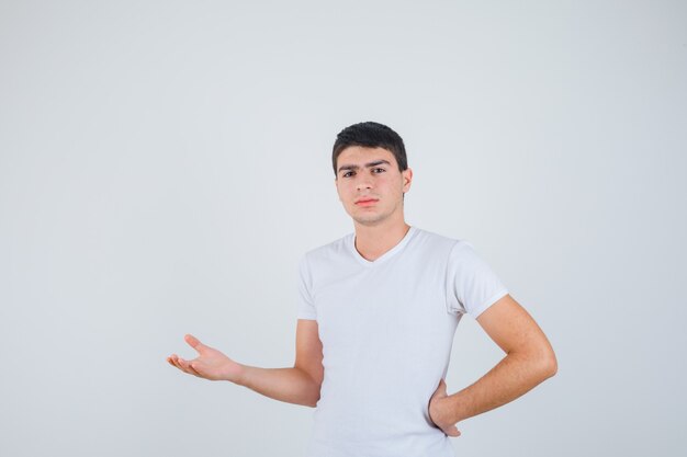 Varón joven fingiendo mostrar algo en camiseta y mirando confiado, vista frontal.