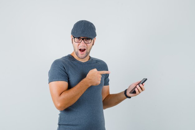 Varón joven en gorra de camiseta gris apuntando al teléfono móvil y mirando asombrado