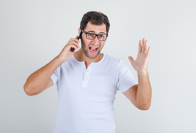 Varón joven gritando por teléfono móvil en camiseta blanca, gafas y mirando irritado