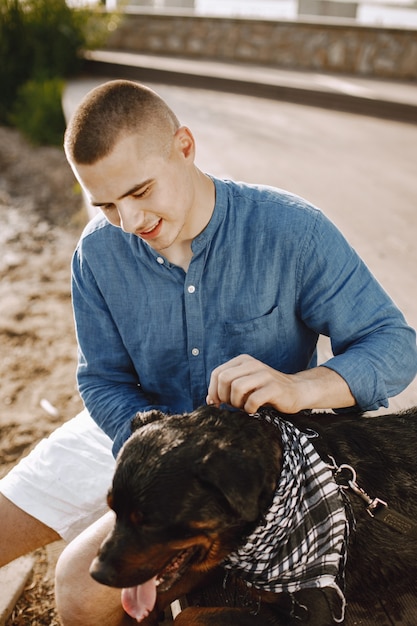 Varón joven guapo en traje casual jugando con lindo perro mientras está sentado cerca del lago. Niño vestido con camisa azul y pantalones cortos de jeans blancos