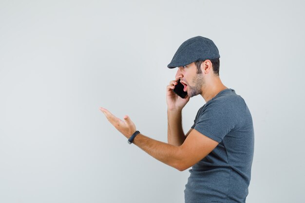 Varón joven hablando por teléfono móvil en la gorra de la camiseta y mirando enojado