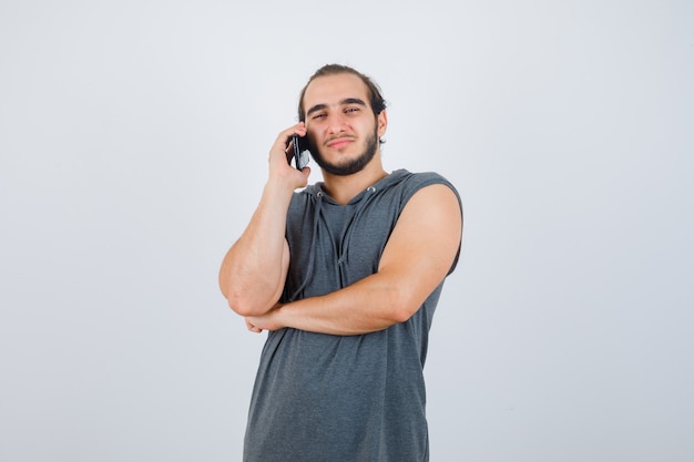 Varón joven hablando por teléfono móvil en sudadera con capucha sin mangas y luciendo guapo. vista frontal.