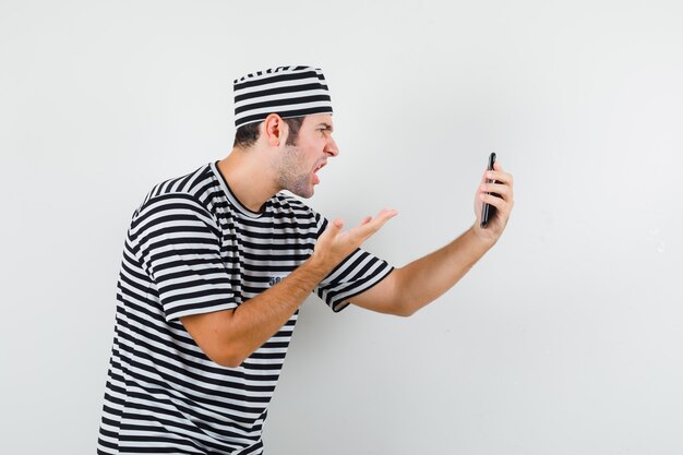 Varón joven hablando por video chat en camiseta, sombrero y con enojo. .