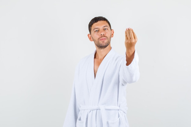 Varón joven haciendo gesto italiano y sonriendo en bata de baño blanca, vista frontal.