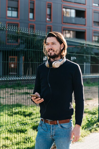 Varón joven hermoso con el teléfono móvil atado en el auricular alrededor de su cuello que se coloca cerca de la cerca