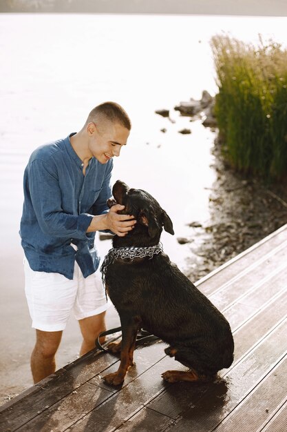 Varón joven hermoso en traje casual que juega con el perro lindo mientras está parado cerca del lago. Niño vestido con camisa azul y pantalones cortos de jeans blancos